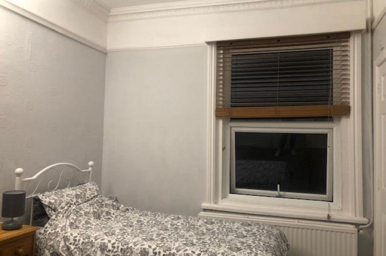 A photo of a bedroom. There is a single bed in the corner of the room with a window on the wall next to it with venetian blinds. Next to the bed is a small bedside table with a lamp on top.