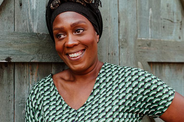 A photo of Karrol - she is a young black female. She is leaning against a wooden door, smiling and looking to the left of the camera