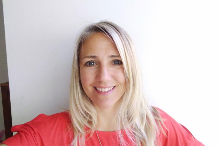 Ellie, a blonde white woman is smiling at the camera. She is wearing a coral top and standing against a white background