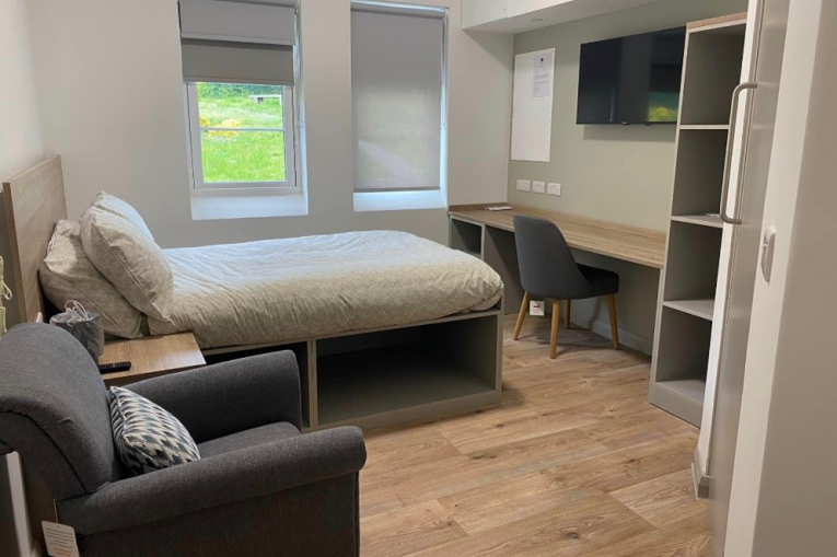 A single bed facing a desk with a TV above it. There is an armchair next to the bed.