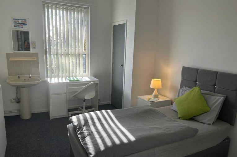 A single bed in a room with a window, bedside table and a small sink