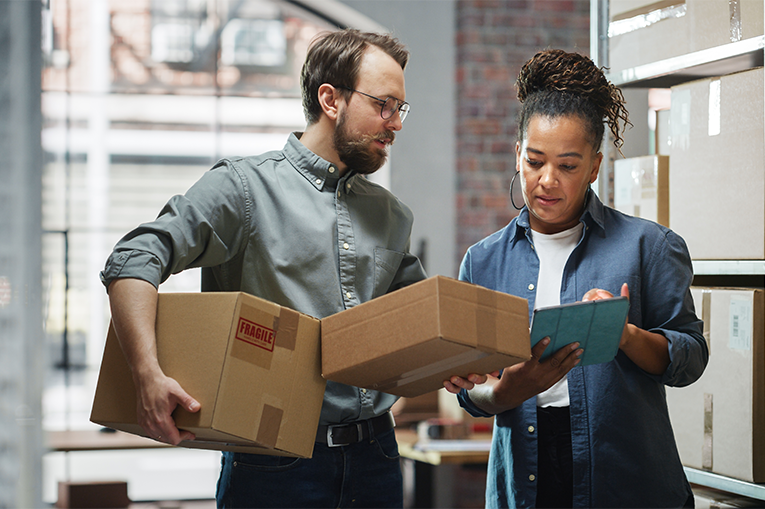 A man and a woman stand together looking at notepad in a brightly lit room. The man is holding cardboard boxes and there are many cardboard boxes stacked behind them in the room.