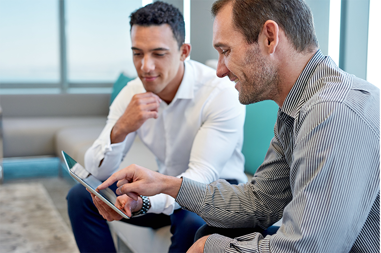Two smartly dressed men are looking at a tablet while sitting on a sofa.