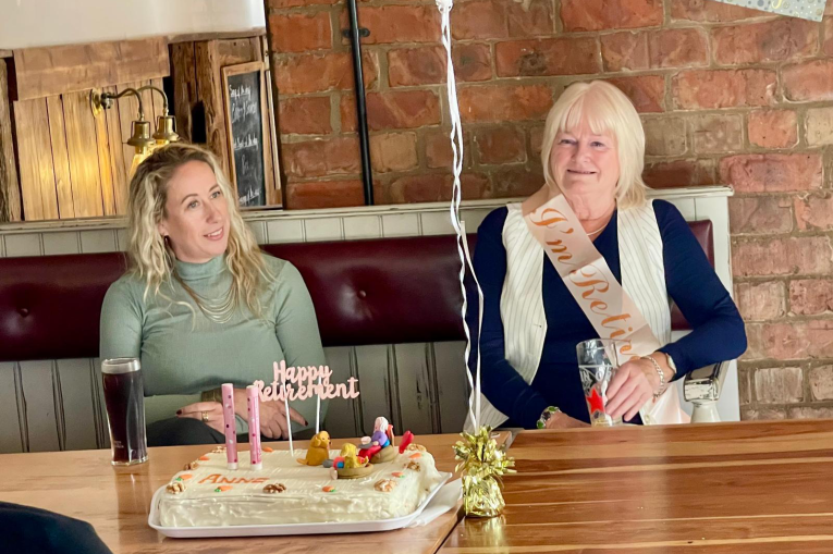 Two women sat at a table with a cake in front of them. The older woman is wearing a sash that says I'm retired