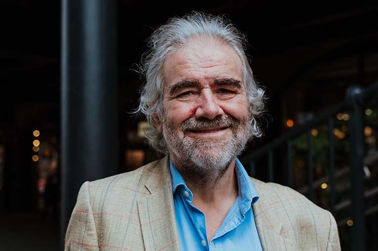 Simon has grey hair and beard and is wearing a blue shirt and light jacket