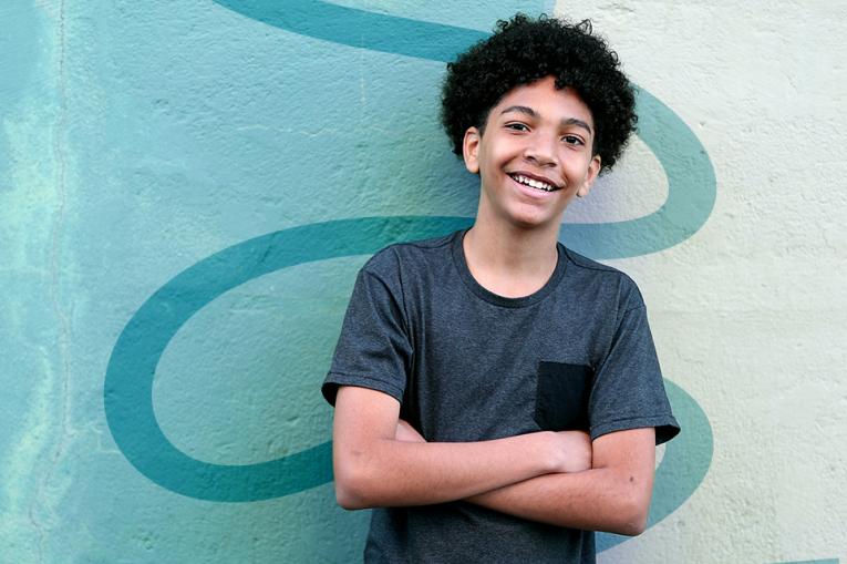 A teenage boy smiles with arms crossed in front of a light blue wall