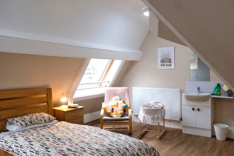 A bedroom with slanted ceilings. There is a single bed with a sink and a Moses basket