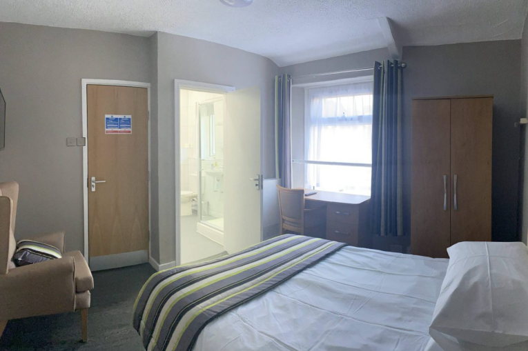 A bedroom with an en suite bathroom. The room contains an arm chair, wardrobe, desk, TV, and bed