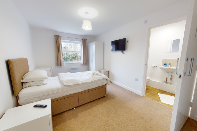 A single bed in a room with a TV mounted on the wall, a bedside table, and an open door to an en-suite bathroom