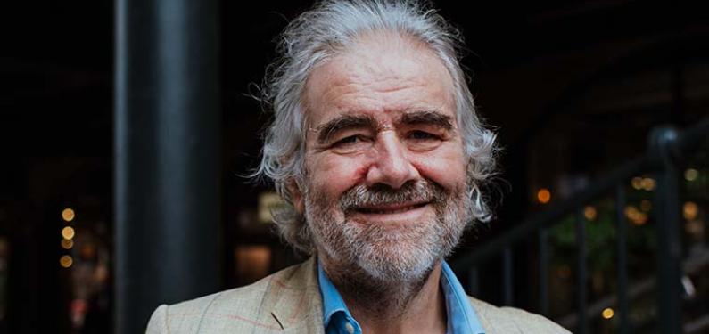 Simon has grey hair and beard and is wearing a blue shirt and light jacket