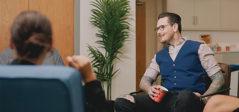 Neil sits in service relaxed drinking a coffee and chatting with colleagues