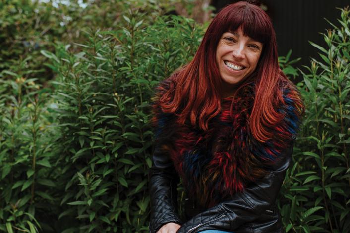 A woman with long dark red hair smiling at the camera sat in front of some bushes