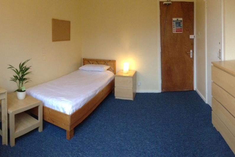 A photo of a bedroom , the bed is in the left hand corner, the door in the right, there is a chest of drawers in the front right corner of the photo. There are blue carpets and magnolia walls 