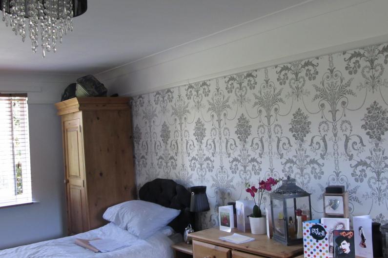 A photo of a bedroom at Allington House. There is an oak wardrobe in the left corner of the room, next to the single bed. There is a chest of drawers on the other side of the bed. The wallpaper is white and grey with a repeating baroque pattern 