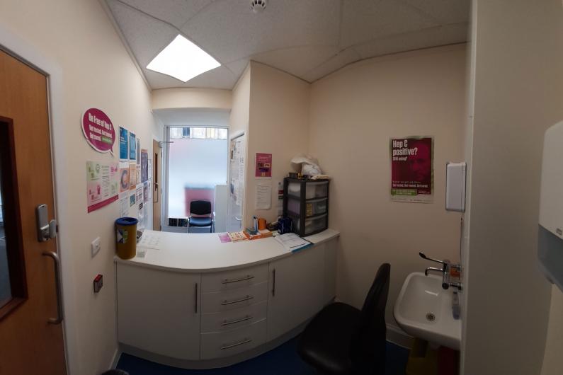 An image of a room, there is a reception desk in the centre of the image, on the left side, there are white walls with posters around them, there is a window at the back of the room