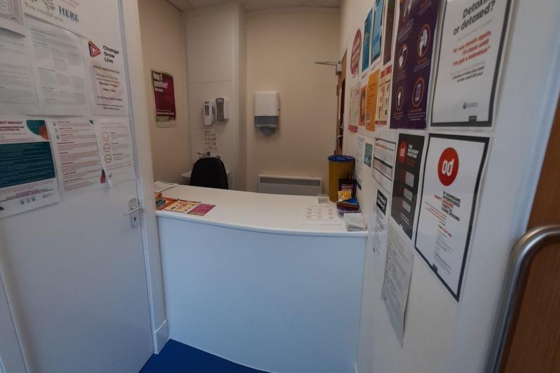 A photo of a reception desk, the desk is in the middle of the room with the two walls either side of it. There is a purple carpet.  