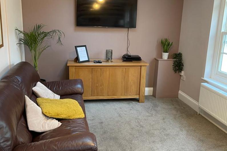 Lounge area - a room with a large brown sofa, a TV mounted on the wall, a storage unit under the tv and a window to the right of the image.