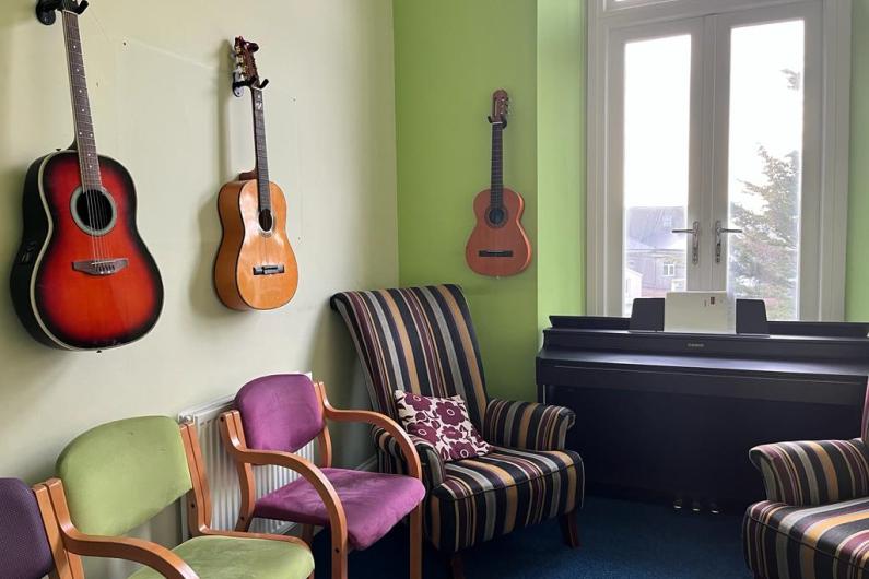 Music room - a room with three brightly coloured armchairs, and an upright piano, a large window. There are two guitars and a ukulele mounted on the wall.