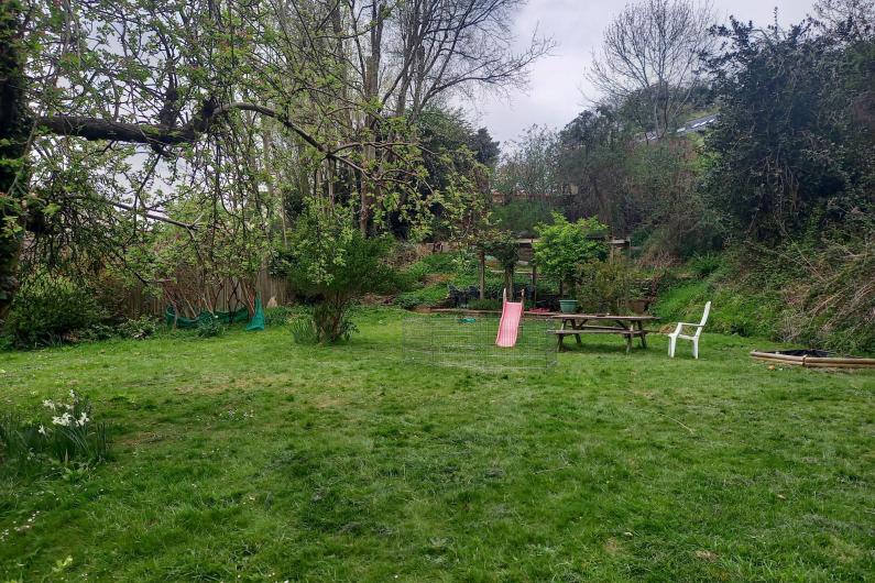 A photo of a big garden. There is plenty of open space and greenery, including grass and trees. There is a child's slide and a wooden picnic bench towards the back of the garden. 