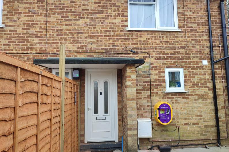 A photo of the outside of a house. It is made of red bricks and it two stories tall. There is a small porch covering the white front door. There are two windows visible on the front of the house. One one the top floor and one one the ground floor. Underneath the ground floor window is a yellow and purple defibrillator box. 