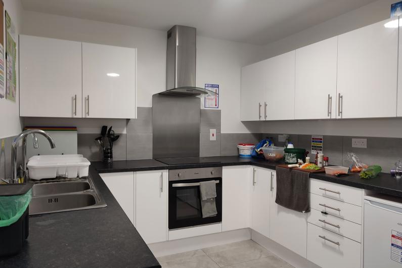 A photo of a kitchen. The cabinets and drawers are all white with silver handles. They have black countertops and a double sink. The over and stove top is on the back wall. There is a salad being prepared on the right hand side.