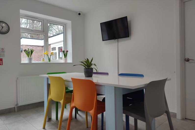 A photo of a white dining area. there is a window on the back wall and a TV on the adjacent wall. A white dining table sits in the middle with rainbow coloured plastic chairs tucked underneath. 