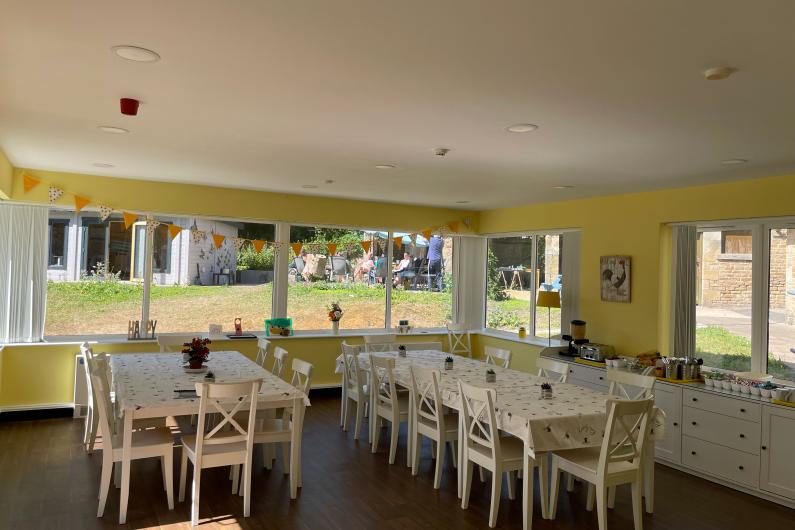 A photo of a dining room. There are two large dining tables, each with wooden chairs around the edge. A counter top runs along one wall with a toaster, appliances and cutlery. A big window wraps around one wall looking out into the garden. The walls are painted a bright Yellow.