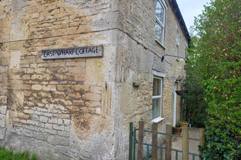 A close up of the corner of an old cottage. There is an old piece drift with the words 'East Wharf Cottage' engraved onto it. There is a small gate and a path leading to a door on the right hand side of the building, which is lined with green bushes. 