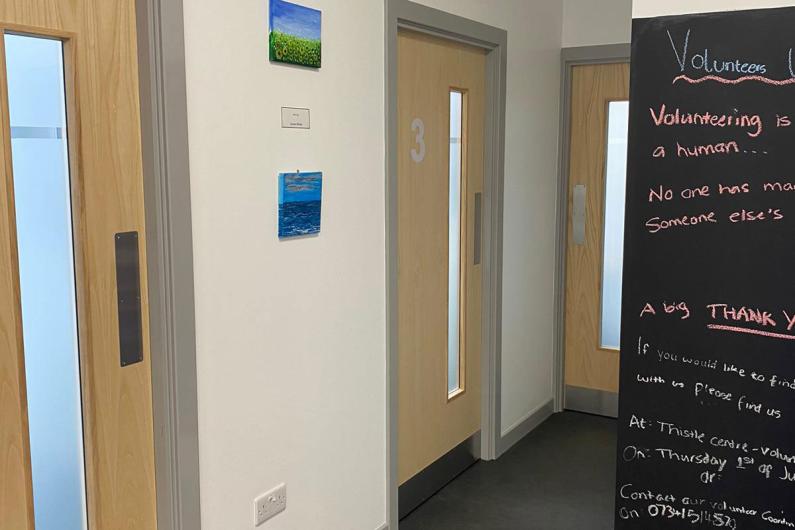 An image of a hallway. There are two doors on the left hand side and one at the end of the hall. On the right hand side, there is a blackboard with writing on it. 