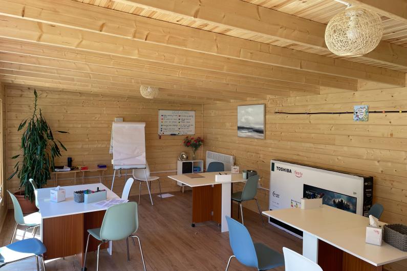 A photo of the inside of a wooden cabin. There are three tables dotted around the room with a few chairs at them. There is a flipchart at the end of the room and a whiteboard on the wall. 