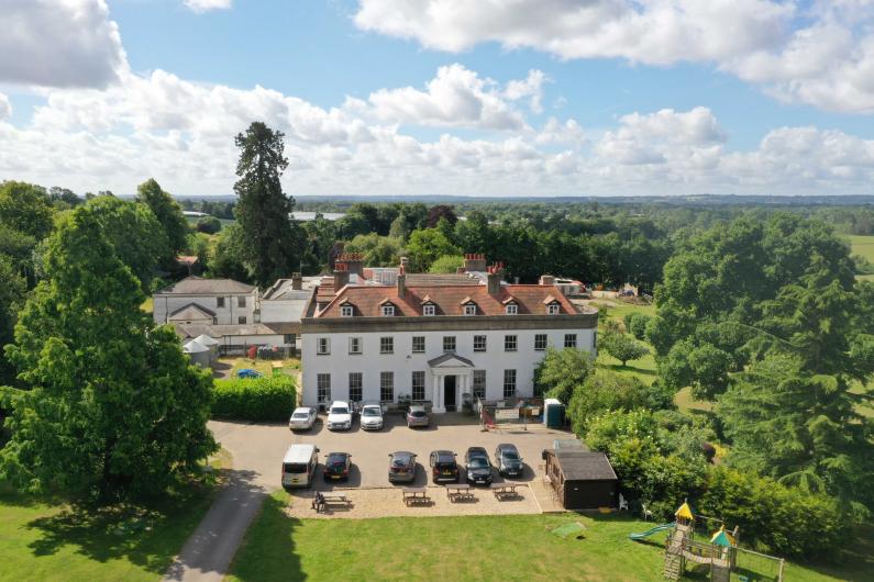 An aerial photographc of Kenworth House on a sunny day. A large white detached stately home with surrounding park. There are cars on a large drive