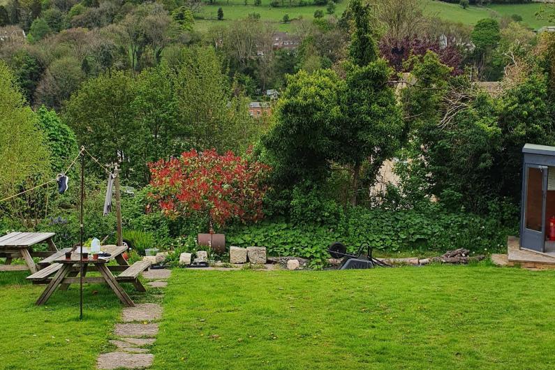 A photo of a garden overlooking rolling hills and fields. There is a small shed just visible at the bottom of the garden in the top right corner. A stone path leads to the bottom of the garden where a washing line is strung up between wooden poles. Two wooden picnic benches are also by the washing line. 
