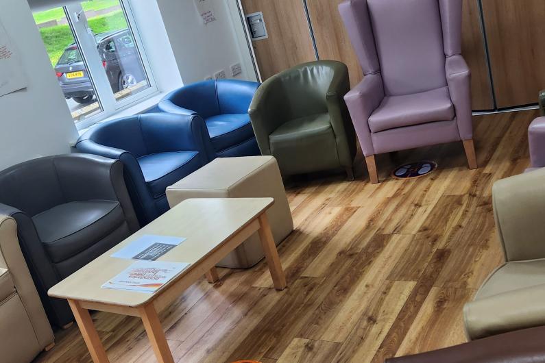 A communal area. there is wooden floors and two windows letting a lot of light in There are a selection of different arm chairs dotted around the edge of the room. 