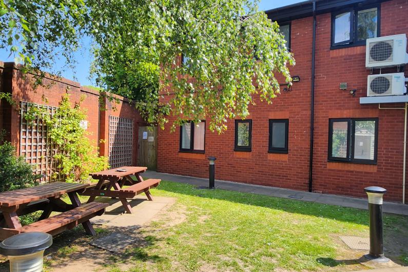 A garden. The Park House building is in the back of the shot with red brick walls. there is a brick wall along the left hand with trellis along the wall. There are two wooden picnic benches underneath a tree.  