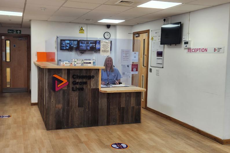 The park house reception area. There is a desk in the middle of the room with perspex glass around it. There is a friendly person sat behind the desk smiling at the camera. 