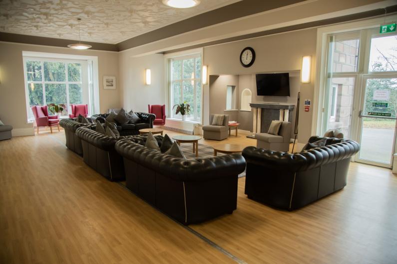 A photo of a living room. there are 5 leather sofas all facing a TV that is mounted on a wall above a fireplace There is a clock on the wall above the TV. In the corner to the room there are arm chairs in front of big windows. 