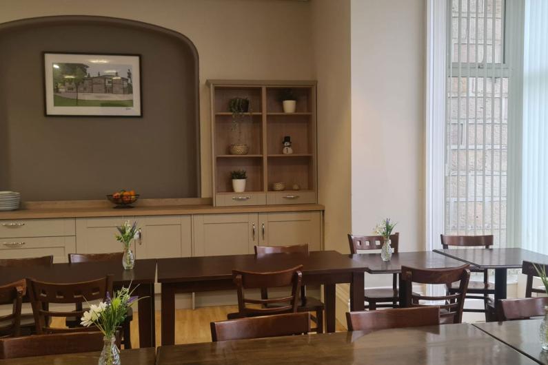 A photo of a dining room. There are 2 rows of dark brown tables with chairs tucked underneath them. There are cupboards and counterspace behind the tables. 