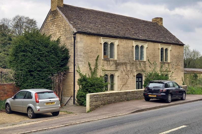 A photo of an old brick house. There are 5 windows on the front of the building along with a door, and a small wall out the front separating it from the public footpath. There are plenty of trees and greenery surrounding the property. 