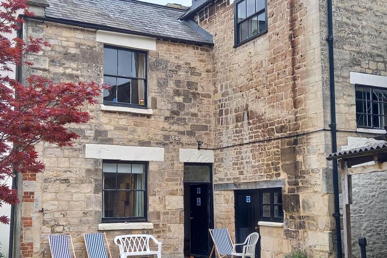 A photo of the courtyard of a house. There are a few windows dotted around the building and a door leading from the house to the courtyard. There are 3 blue and white striped deck chairs in the garden along with a plastic garden chair and two person plastic garden bench. 