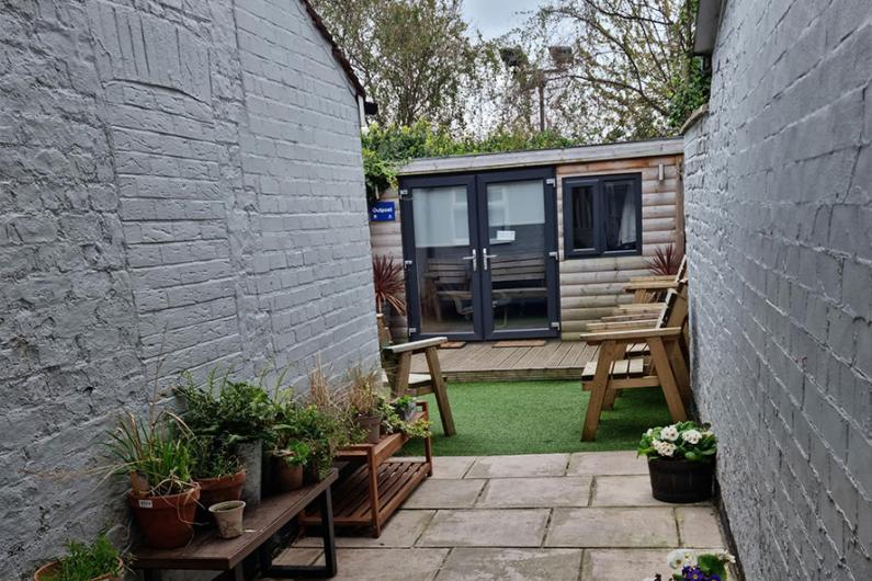 A Photo of a courtyard. There's a patio in the foreground, white walls either side, some grass and a garden office at the far end