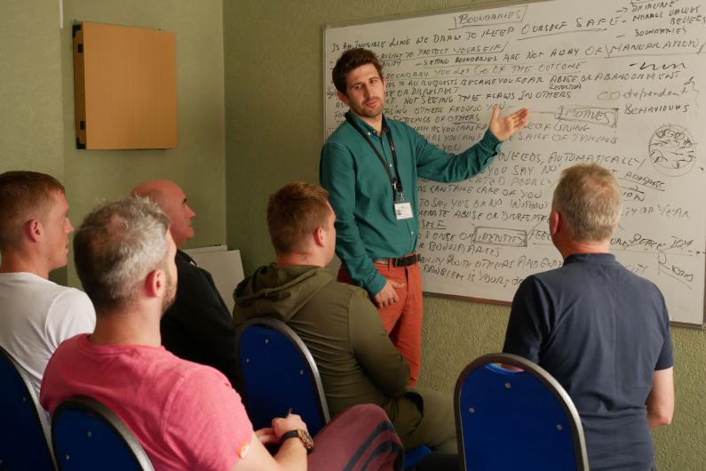 A group therapy class - with a white man standing at a whiteboard and a group of five men on chairs facing towards him