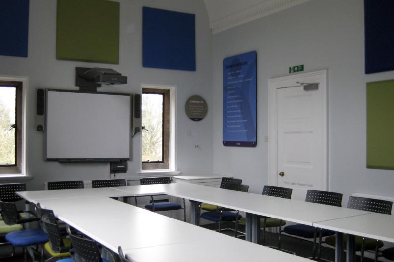 Yeldall Manor - group room. Tables form a U shape with chairs surrounding them. There is a whiteboard, windows and large colourful panels on the walls
