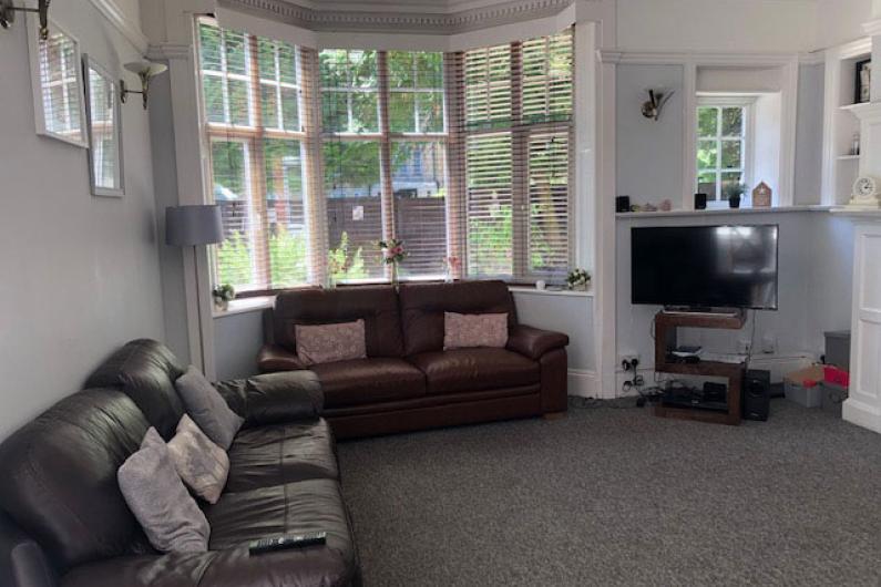 A photo of a communal area. There is a bay window along the back wall with venetian blinds. In front of the window is a two seater leather sofa, with another two seater leather sofa next to it. There is a TV in the corner of the room. 