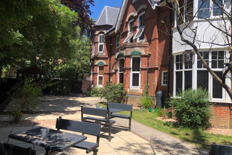 A photo of a garden in front of the red brick building. There is a patio with multiple tables and chairs and plenty of trees and bushes around the edge.