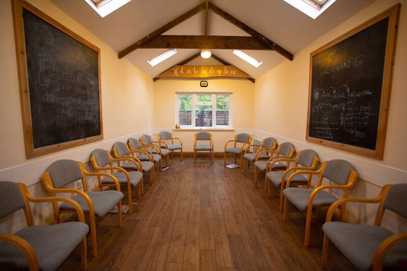 A photo of a long room with a window on the back wall. there are wooden beams lining the ceiling, a blackboard on each wall and grey chairs around the edge of the room. 