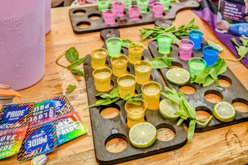 A photo of a woman sat behind a table. On the table is a selection of shot glasses