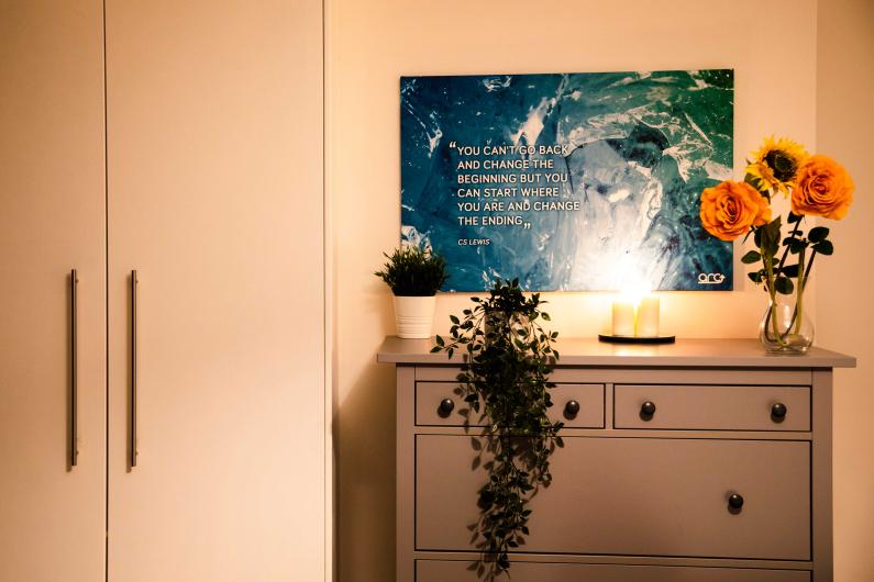  a chest of drawers in the bedroom next to a wardrobe. On the chest of drawers are some fake flowers and candles. there is a piece of art on the wall. 