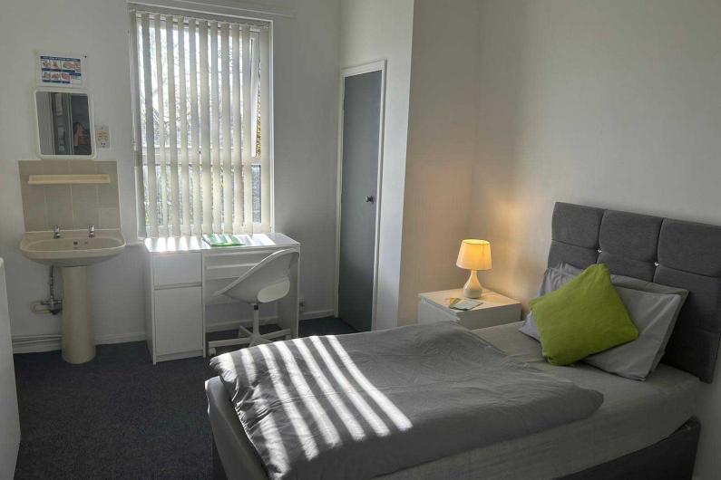 A single bed in a room with a window, bedside table and a small sink