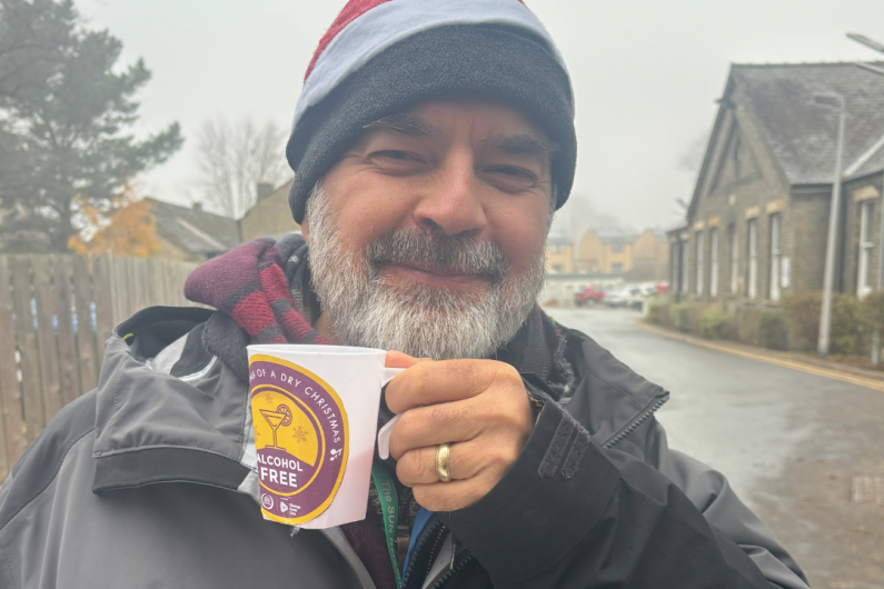 A man in a woolly hat and thick coat holding a mug up to the camera