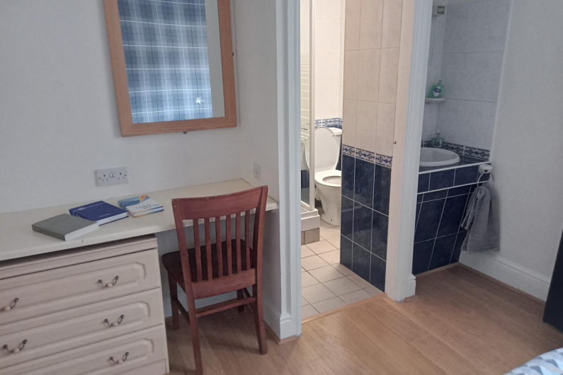A desk  is in the corner of the room next to a door that leads to a bathroom. On the other side of the doorframe is a sink basin.